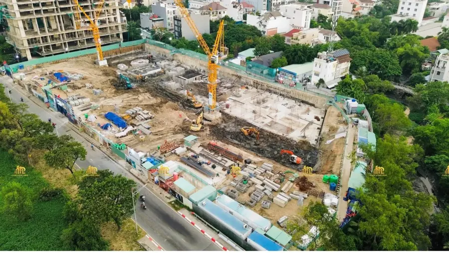Ảnh chụp từ trên cao: hố móng đã đào, hai cần cẩu tháp dựng trong công trường, khu dân cư hiện hữu bao quanh