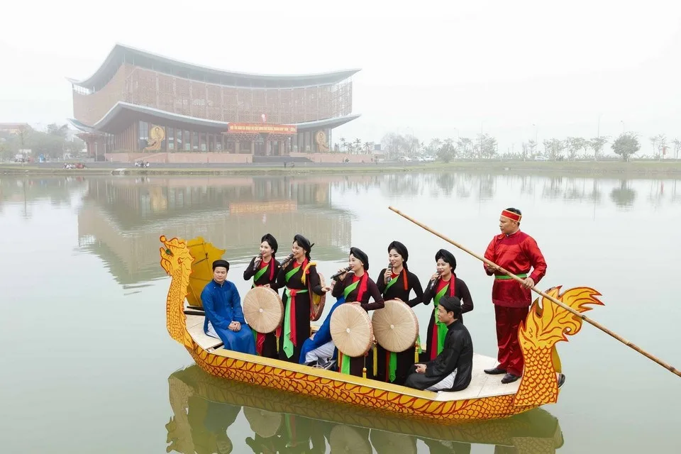 Các liền anh liền chị hát Quan họ trên thuyền rồng giữa mặt nước, phía sau là mái đình cong